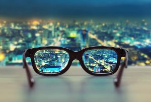 A pair of black eyeglasses sits on a flat surface, their lenses drawing attention to a sharp cityscape at night. Detailed buildings and lights are clearly visible, while the surrounding city is blurred, emphasizing the remarkable clarity provided by the glasses.