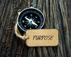A compass with a rustic string loop rests on a textured wooden surface, drawing attention to its vintage charm. Beside it lies a brown tag with the word "PURPOSE" handwritten in black ink, symbolizing direction and meaning in life.