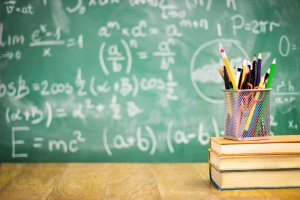 A chalkboard filled with math equations serves as the background. In the foreground, a metal mesh container holds various pencils and pens, resting on top of two stacked books on a wooden desk, creating an atmosphere that feels quintessentially teacher-like.