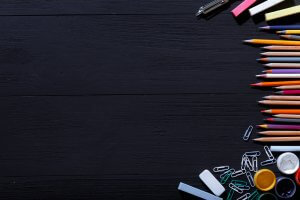 A dark wooden surface with an assortment of stationery along the edges, including colored pencils, erasers, paper clips, binder clips, a pencil sharpener, and other school supplies that any teacher would need. The center of the image is empty, creating a contrast against the vibrant items.