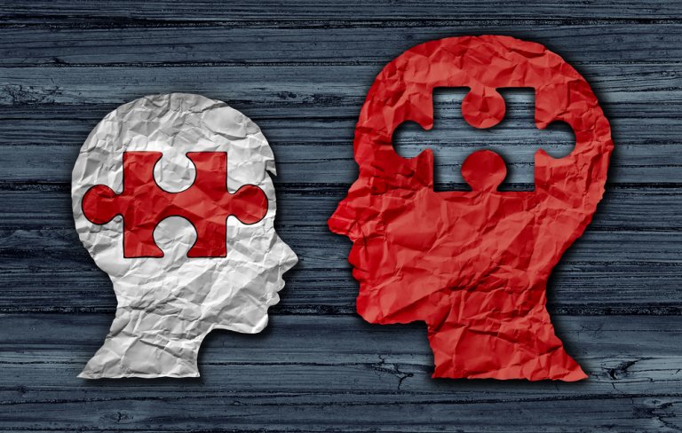 Illustration of two crumpled, paper-cut profiles: a small white head with a missing puzzle piece forming its brain and a large red head with a puzzle piece-shaped cutout. They face each other like a teacher and student, set against a textured wooden background.