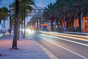 A city street at dusk with palm trees lining the sidewalk, blurred car lights creating light trails, and illuminated shops including a Crocs store in the background. A few blurry pedestrians walk along the sidewalk.