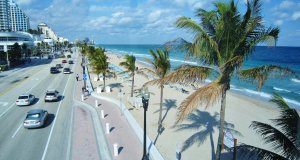 A coastal road runs parallel to a sandy beach lined with tall palm trees. Cars drive on the road, and beachgoers relax under umbrellas on the beach. The blue ocean waves gently crash against the shore under a partly cloudy sky.