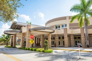 A modern building with a curved entrance canopy, surrounded by palm trees. The structure features large windows and columns. A stop sign and pedestrian crossing sign are visible near the entrance. The sky is clear with a few fluffy clouds.