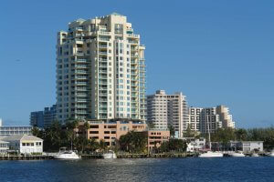 A cluster of high-rise apartment buildings overlooks a marina filled with various boats on a sunny day. The buildings are situated along the edge of a waterfront, with clear blue skies and calm water enhancing the serene atmosphere.