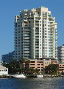 A tall, modern apartment building with numerous balconies stands beside a waterway. Palm trees and a few boats are docked near the building. The sky is clear and blue, suggesting a sunny day. Other buildings can be seen in the background.