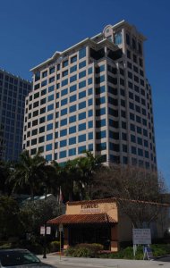 A tall, multi-story office building stands against a clear blue sky. In front, there is a smaller building with a sign that reads "Edible Arrangements Flowers and Gourmet Chocolate." Several palm trees and a signboard with contact information are visible.