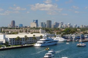A vibrant harbor scene with multiple yachts and boats docked in the water. Modern city skyscrapers and various buildings stand tall in the background. The sky is clear with a few white clouds, creating a picturesque view of an urban waterfront.