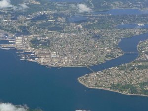 Aerial view of a coastal city with dense buildings, roads, and several bridges connecting land over blue water; patches of greenery and clouds are visible above and around the area.