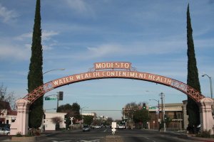 A large archway sign over a city street reads “MODESTO WATER WEALTH CONTENTMENT HEALTH,” flanked by two tall cypress trees, with buildings and traffic lights visible in the background.