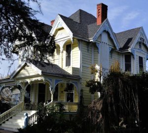 A large, two-story Victorian-style house with yellow siding, white trim, decorative woodwork, a covered porch, and red brick chimneys, partially shaded by trees and surrounded by greenery.