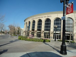 A wide view of the Gallo Center for the Arts in Modesto, California. The building features a curved facade with tall, arched windows. The street in front is decorated with bare trees and street lamps, and there is a red banner hanging from a lamp post.