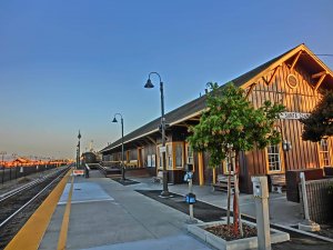 A wooden train station with a sign reading "Santa Clara," a tree in front, lampposts along the platform, and train tracks extending into the distance under a clear blue sky.