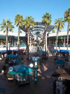 Outdoor shopping mall with palm trees, blue awnings, and vendor kiosks along a wide walkway. Stairs lead to a second level with more shops. The sky is clear and sunny.