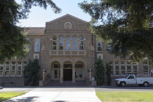 A historic brick building with arched windows, a decorative façade, and a central entrance with steps. Tall trees frame the building, and a white pickup truck is parked to the right.