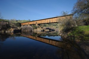 A long, wooden covered bridge spans over a calm, reflective river, set against a clear blue sky. The bridge and surrounding trees are mirrored in the water below. The scene is pastoral with green hills and sparse vegetation.