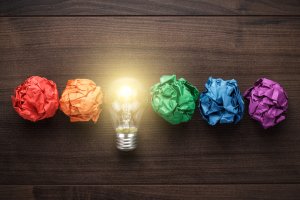 A glowing lightbulb is flanked by crumpled paper balls arranged in rainbow order: red, orange, yellow, green, blue, and purple. Set against a dark wooden background, the scene symbolizes creativity and ideas sparking to life—like the innovative breakthroughs often credited to billionaires.