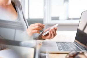 A person is sitting at a desk with a laptop open in front of them, holding and using a smartphone. The scene is well-lit, with blurred elements of the workspace visible, indicating a modern, professional environment perfect for seeking out career advice.