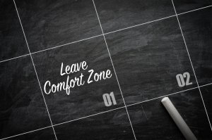 A chalkboard calendar with the words "Leave Comfort Zone" written in one of the squares. There is a piece of chalk resting near the corner of the square labeled "02," urging a break from complacency.