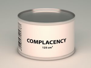 A silver can adorned with a plain light-colored label stands against a neutral background. The label boldly reads "COMPLACENCY" in black letters, with "125 cm³" beneath it. Part of a barcode is visible on the left side of the can, perfectly encapsulating the essence of complacency.