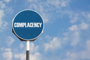A blue road sign with the word "COMPLACENCY" boldly written in white uppercase letters stands out against a backdrop of a clear sky with scattered clouds. The sign is firmly mounted on a pole, serving as a stark reminder.