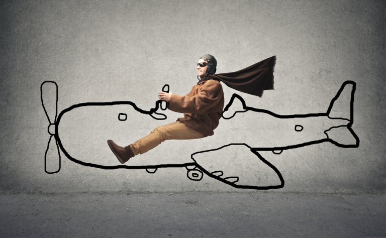 A person wearing a pilot's helmet, goggles, scarf, brown jacket, and pants is positioned as if sitting in and flying a hand-drawn airplane on a wall. The scene integrates the person's body into the drawing, creating a whimsical depiction of their dream job with imaginative flair.
