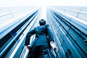 A person in a business suit stands between two tall, modern glass skyscrapers, looking up towards the sky. The perspective emphasizes the height and grandeur of the buildings. The scene conveys a sense of ambition and aspiration, as if they are envisioning their dream job within these towering structures.