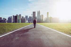 A person in a suit strides toward a distant city skyline on an empty road. The skyline features various skyscrapers, and the road is flanked by green fields, with the bright sun casting light from the right, creating a serene contrast of nature and urban life—an inspiring journey towards their dream job.