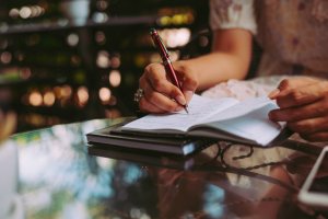 Close-up of a person writing in a notebook with a pen, seated at an outdoor glass-top table. The setting resembles a cafe or garden event, with blurred greenery in the background. The writer, wearing a ring and a light-colored top, seems focused on their notes.