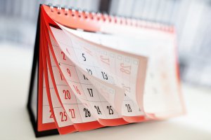 Close-up of a desk calendar with red and black numbers. Pages are flipping, showing dates from various months, each potential event waiting to be marked. The calendar has a spiral binding at the top and is set against a blurred background. The days and dates are visible, although slightly blurred due to the motion.