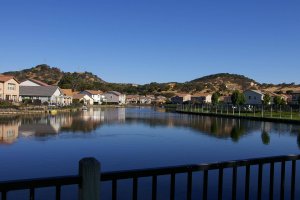 A calm lake reflects rows of houses and trees under a clear blue sky, with hills in the background and a black railing in the foreground.