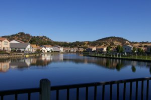 A serene lakeside community featuring houses along the water's edge, with clear blue skies overhead. Hills with light vegetation rise in the background. A fence in the foreground spans the scene, reflecting in the calm water below.