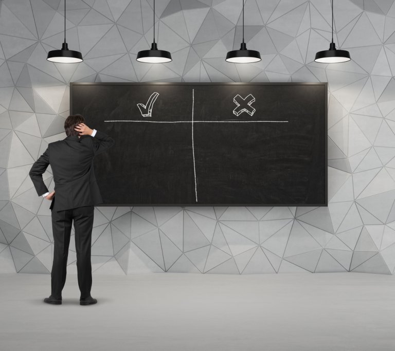 A person in a suit stands in front of a large blackboard divided into four sections. One section has a checkmark, while another has an X. Possibly contemplating corporate hiring decisions, they have their hand on their head. The room is modern, with geometric wall patterns and hanging lights.
