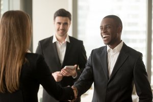 Two business professionals are shaking hands, one man and one woman, both dressed in suits. A third professional, possibly an employer, stands behind them smiling and holding a tablet. The setting appears to be a bright office with windows in the background.