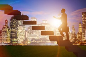 A person in silhouette walks up floating steps that ascend into the sky against the backdrop of a city skyline at sunset, with the sun glowing brightly behind the buildings. The scene, reminiscent of an ambitious climb one makes in an employer's world, conveys both aspiration and progress.