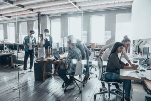 A modern open-plan office with several people working at desks equipped with computers. In this vibrant public sector workspace, a few individuals are standing and discussing near glass walls, while others are seated and focused on their tasks. Tall windows let in natural light.