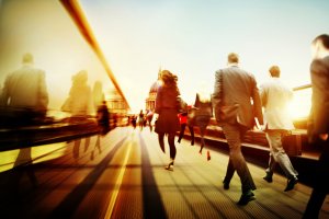 A dynamic city scene at sunset shows a crowd of people in business attire, including many from the public sector, walking across a bridge. The sun casts long shadows while the blurred motion captures the fast-paced movement of commuters against a backdrop of a domed building in the distance.