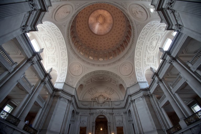A grand architectural interior featuring an ornate, domed ceiling with intricate patterns and designs. Tall columns line the sides, with archways and balconies above. The overall design is elaborate, showcasing classical architectural elements and detailed craftsmanship often seen in public sector buildings.