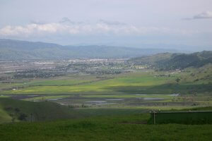 A wide-angle view of a verdant valley with patches of green fields and streams under a cloudy sky. The landscape is bordered by rolling hills and distant mountains. Small clusters of buildings are visible in the middle ground.