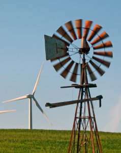 A landscape image shows a traditional windmill in the foreground with rusty metal blades, and a modern white wind turbine in the background. Both structures stand on a grassy field under a clear blue sky.