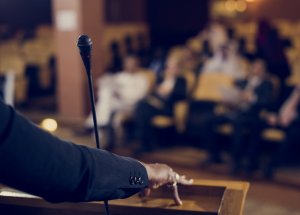 A speaker at a podium is addressing the audience through a microphone, with the focus on the microphone and their arm, while the seated audience appears blurred in the background.