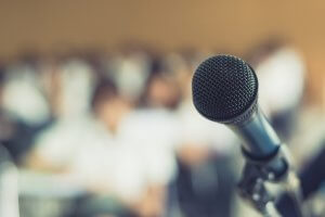 A close-up of a microphone with a blurred background, suggesting a crowd or audience. The focus is on the microphone, with the figures in the background appearing indistinct, implying that the scene is set for a speaker's speech, performance, or presentation.