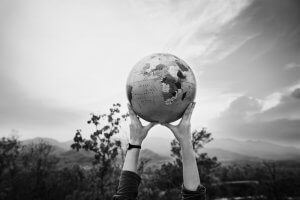 Black and white image of two hands holding up a globe, symbolizing global awareness and exploration. The backdrop of trees, a cloudy sky, and mountainous terrain frames the scene outdoors. This could easily be seen as an employer's commitment to sustainability and worldwide responsibility.