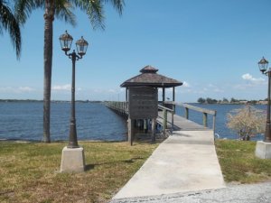 A pathway with street lamps on each side leads to a wooden dock extending over a calm body of water. A palm tree stands to the left of the path. In the distance, a clear blue sky is visible above the water.