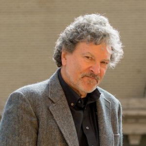 A man with curly gray hair and a beard is wearing a gray tweed blazer over a black shirt. He has a slight smile, radiating perfection, and is standing outside against a beige brick wall background. The lighting suggests a sunny day.