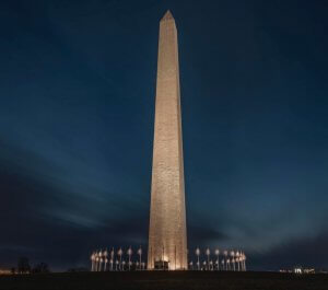 Nighttime view of the illuminated Washington Monument, standing tall against a dark blue sky. The surrounding flagpoles are slightly blurred due to long exposure, highlighting the monument's grandeur and prominence. Lights at the base add a warm glow, reminiscent of preparation for a federal job interview.