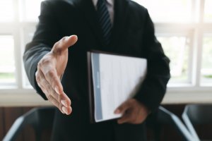 A person dressed in a suit extends their right hand for a handshake while holding a folder or document in their left hand, ready for the job! The background features large windows with natural light.