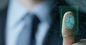 A close-up of a person using biometric technology. The person is pressing their thumb on a translucent screen, displaying a detailed digital fingerprint and technological overlay. The background features a blurred, formal outfit, hinting at a professional setting with strict adherence to HIPAA regulations.