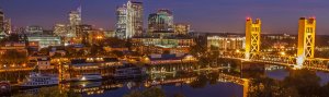 Sacramento cityscape at dusk featuring illuminated buildings and the iconic Tower Bridge reflecting on the calm waters of the Sacramento River. The sky is transitioning from twilight to night, casting a serene ambiance over the scene.