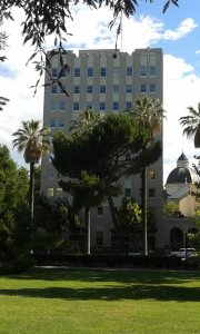 A tall, beige office building with many windows stands behind palm trees and greenery, with a domed building partially visible to the right under a partly cloudy sky.